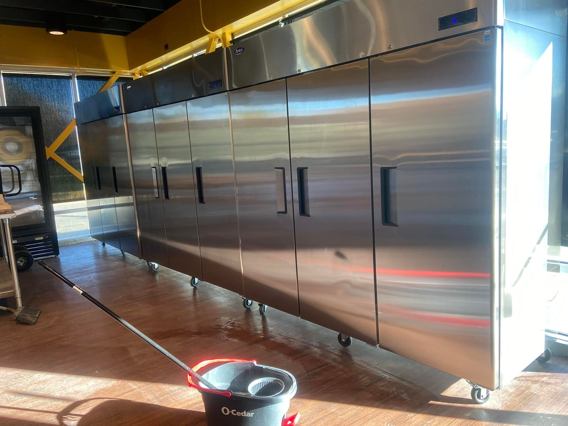 Commercial stainless steel refrigerators in a kitchen, with a mop and bucket in front.