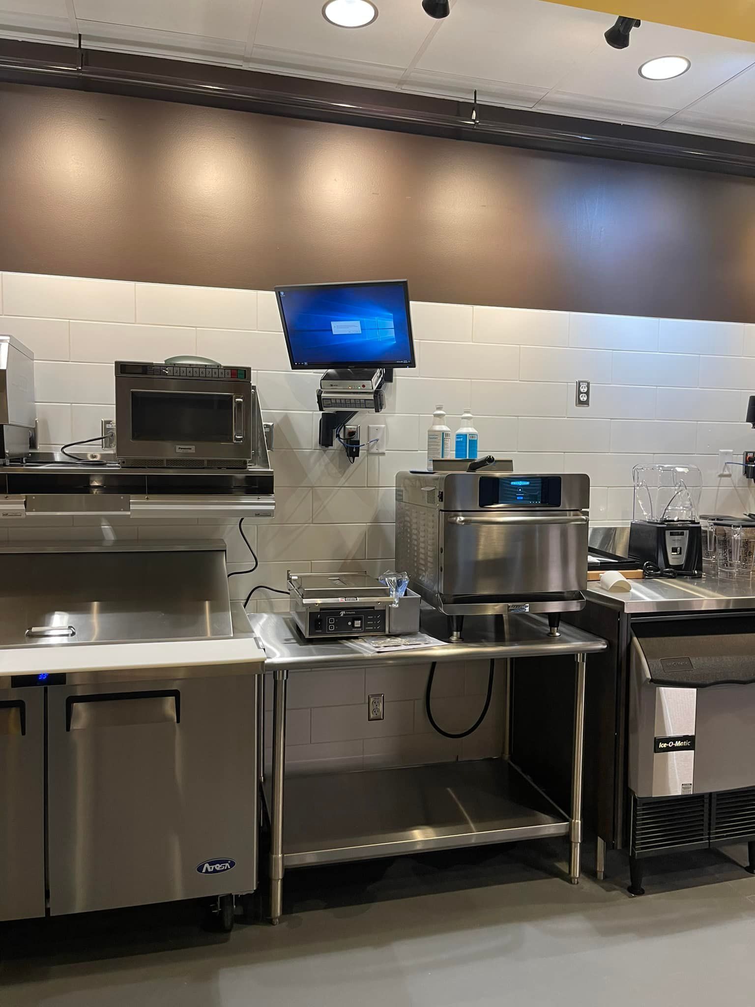 Commercial kitchen with stainless steel appliances, microwave, oven, and mounted monitor.