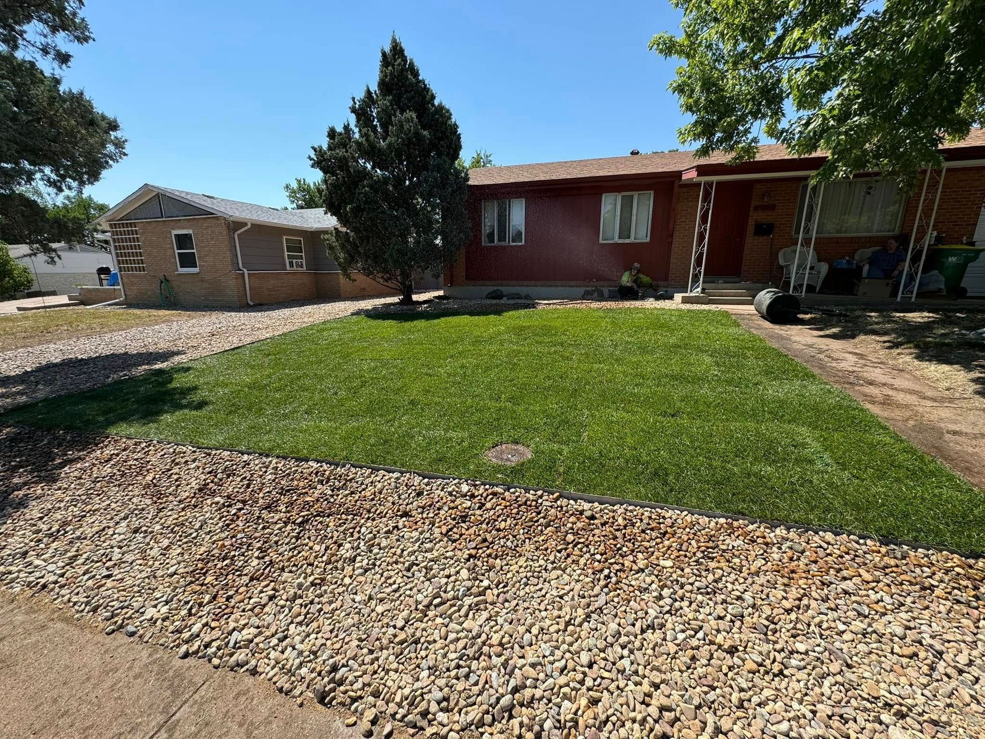 A house with a green lawn and rock border.  A small tree stands in the yard. Blue sky.