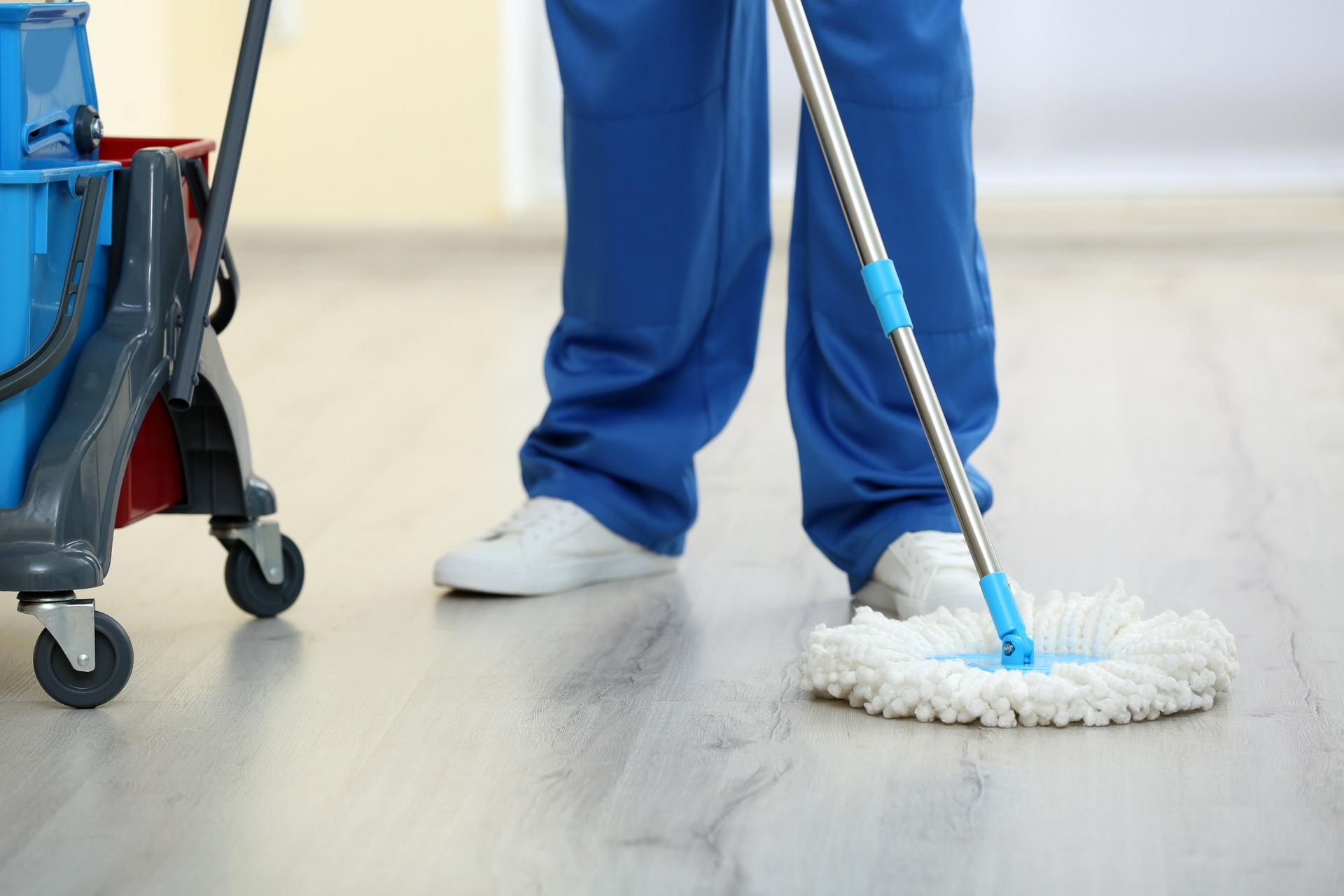 Person in blue uniform mops a light wood floor with a blue mop; cleaning cart on the left.