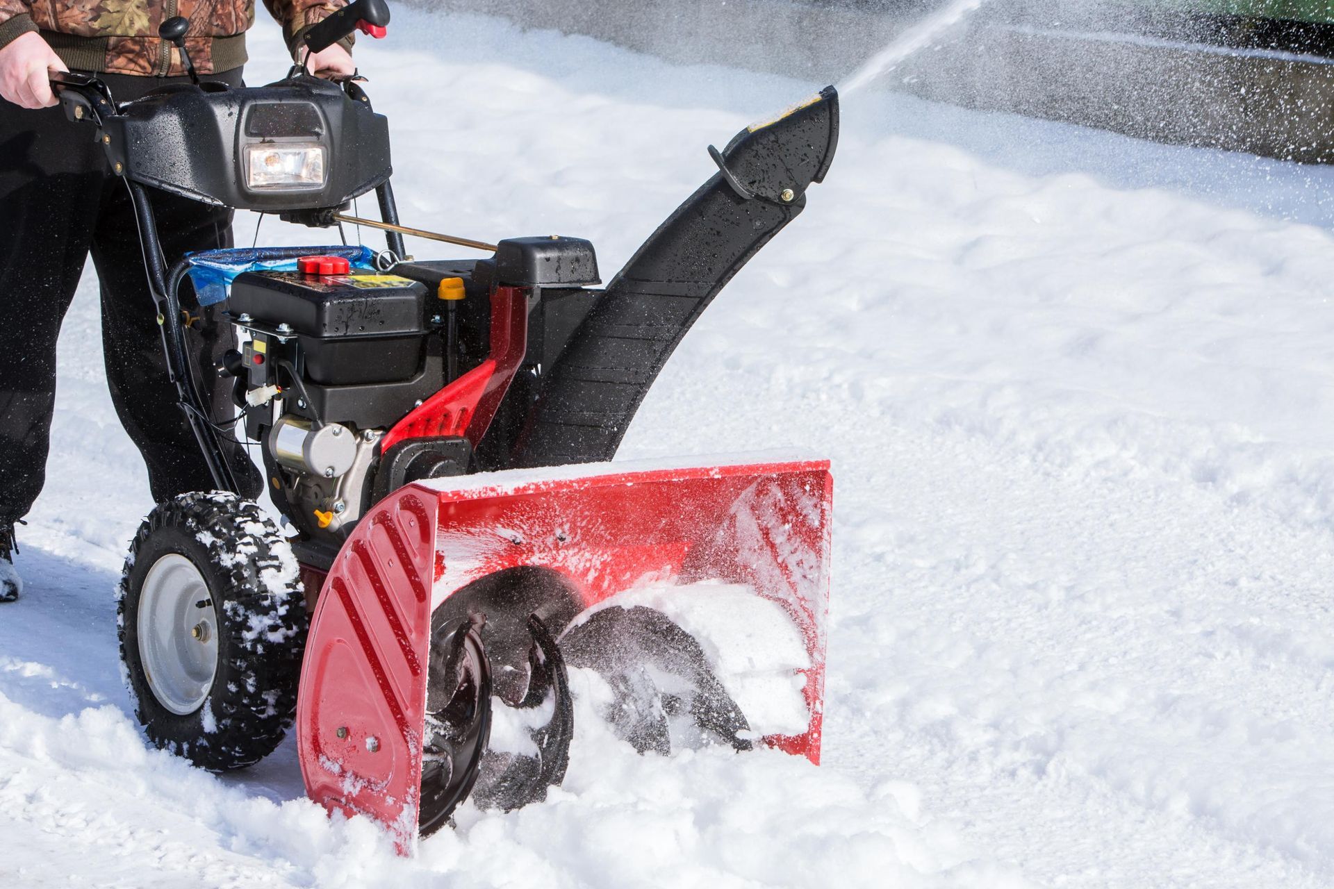 Person operating a red and black snowblower, clearing snow from a driveway.