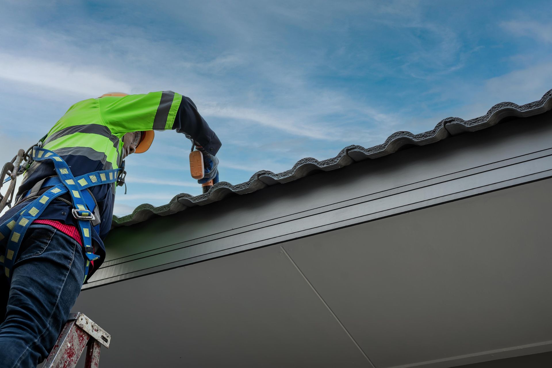 Construction worker on a roof using a drill, wearing a safety harness and high-visibility vest. Blue sky background.