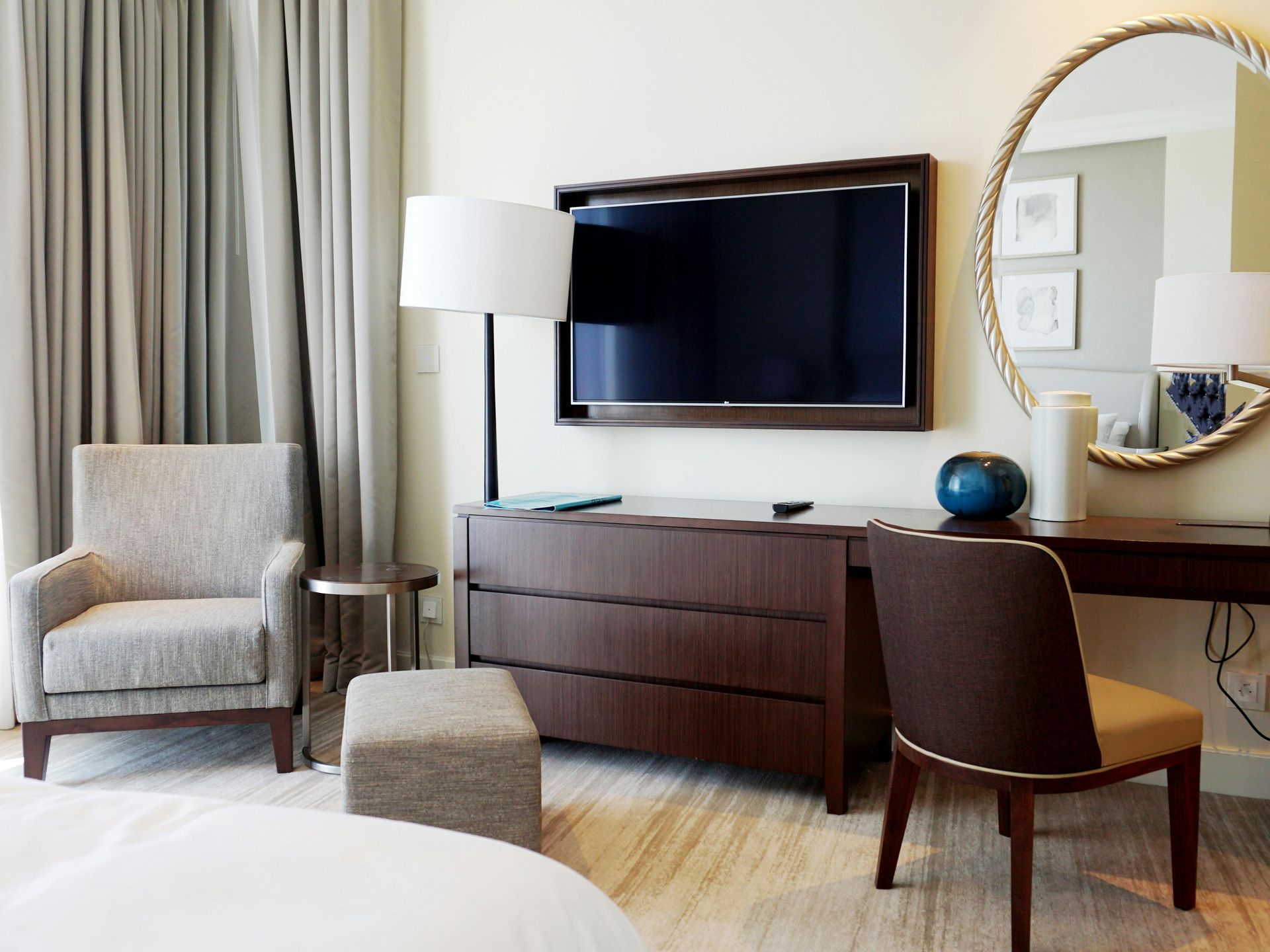 An image of hotel room with a TV, mirror, lamp, chairs and wooden dresser. 