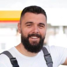 Homem de barba escura sorri, vestindo camisa branca e macacão, em pé do lado de fora.