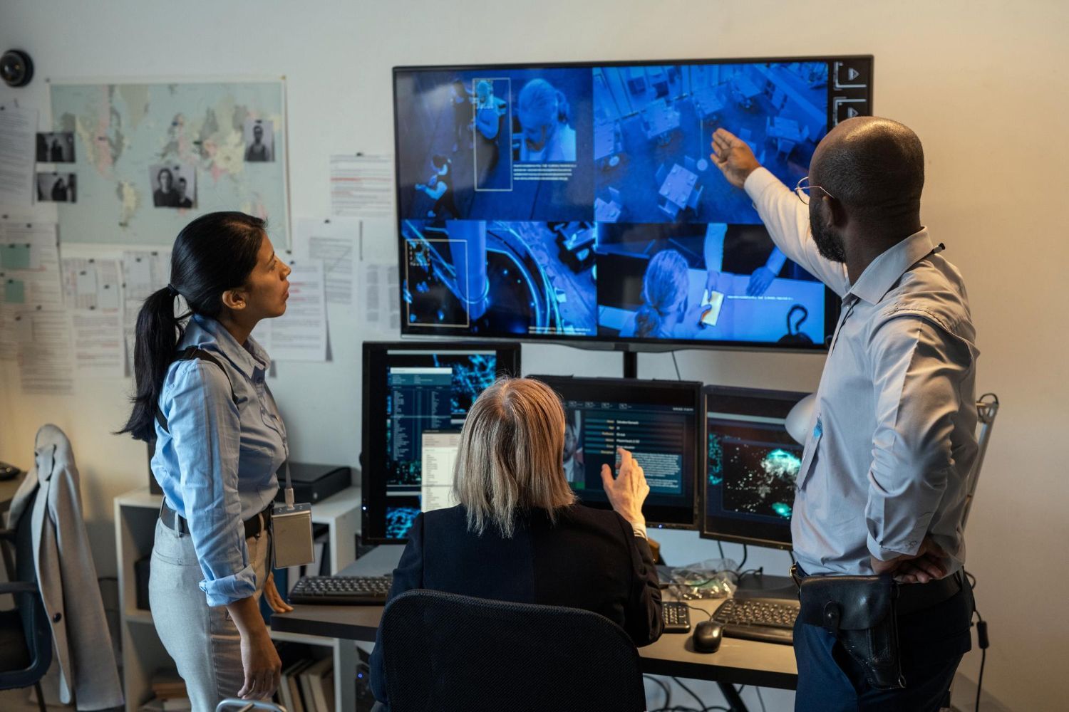 Três pessoas em uma sala de controle, analisando imagens de vigilância em várias telas. 