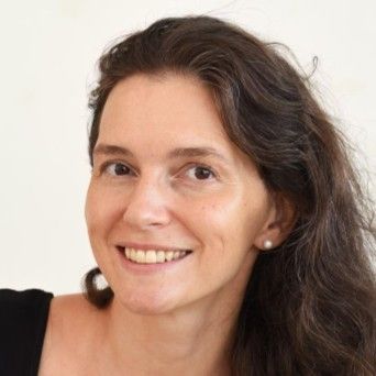 Mulher sorridente, cabelos escuros, vestindo uma blusa preta, brinco de pérola pequeno, contra um fundo claro.