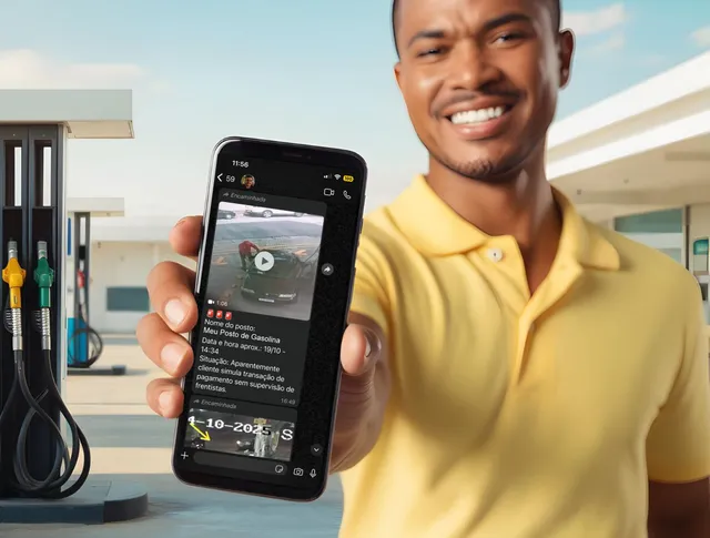 Homem de camisa amarela sorrindo, segurando um celular que exibe um vídeo; posto de gasolina ao fundo.