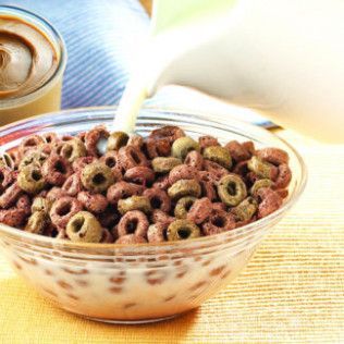 Milk being poured into a bowl of chocolate cereal rings; glass bowl, mug in background.