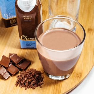 Chocolate protein drink in a glass with chocolate pieces and a carton on a wooden board.