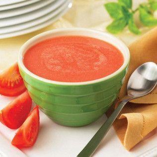 Green bowl of tomato soup, with sliced tomatoes and a spoon on a white plate.