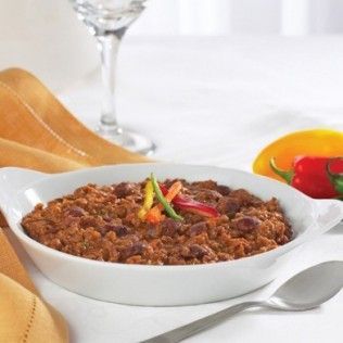 Chili in white bowl, garnished with peppers, on a white table. A spoon and wine glass are nearby.