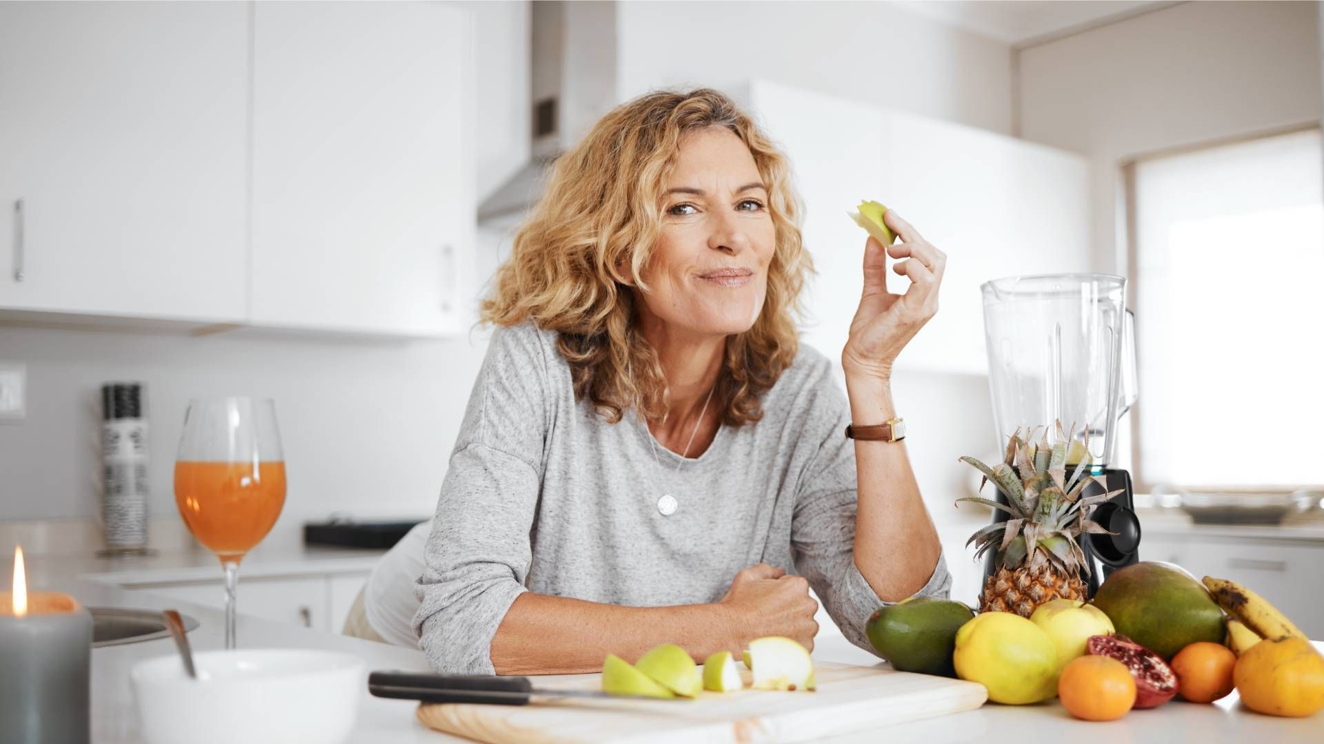 Woman in kitchen holding lime, surrounded by fruits and smoothie ingredients.