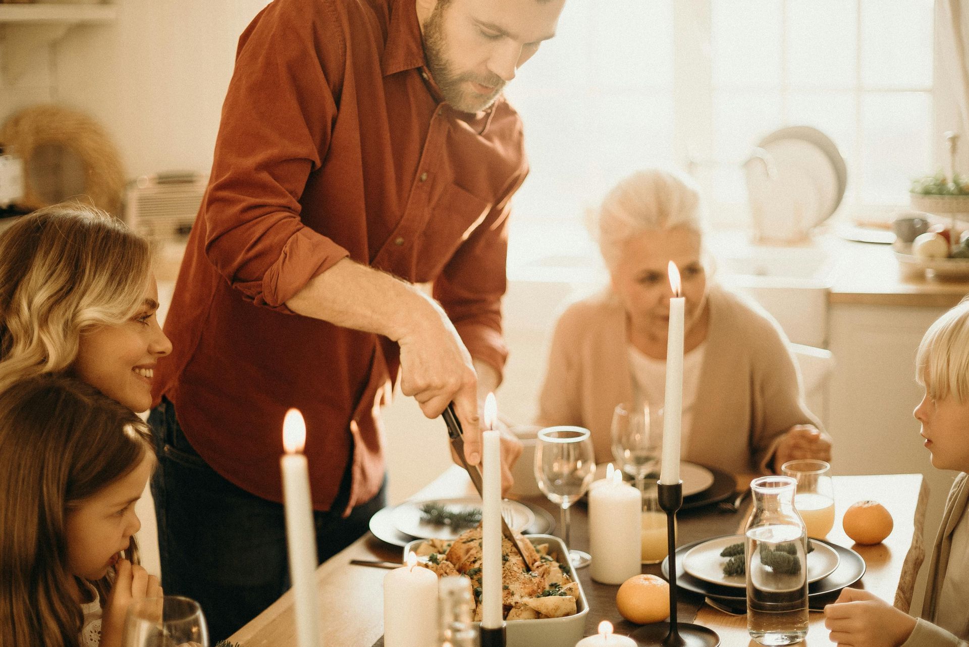 Man carves food at a table. Family watches with smiles; candles and food on the table.