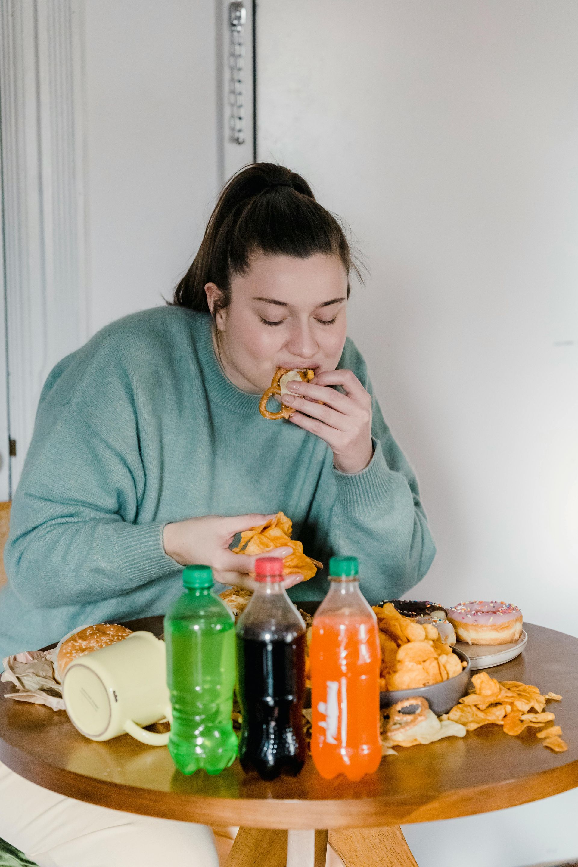 Woman eating a hamburger, surrounded by junk food and soda bottles on a table.