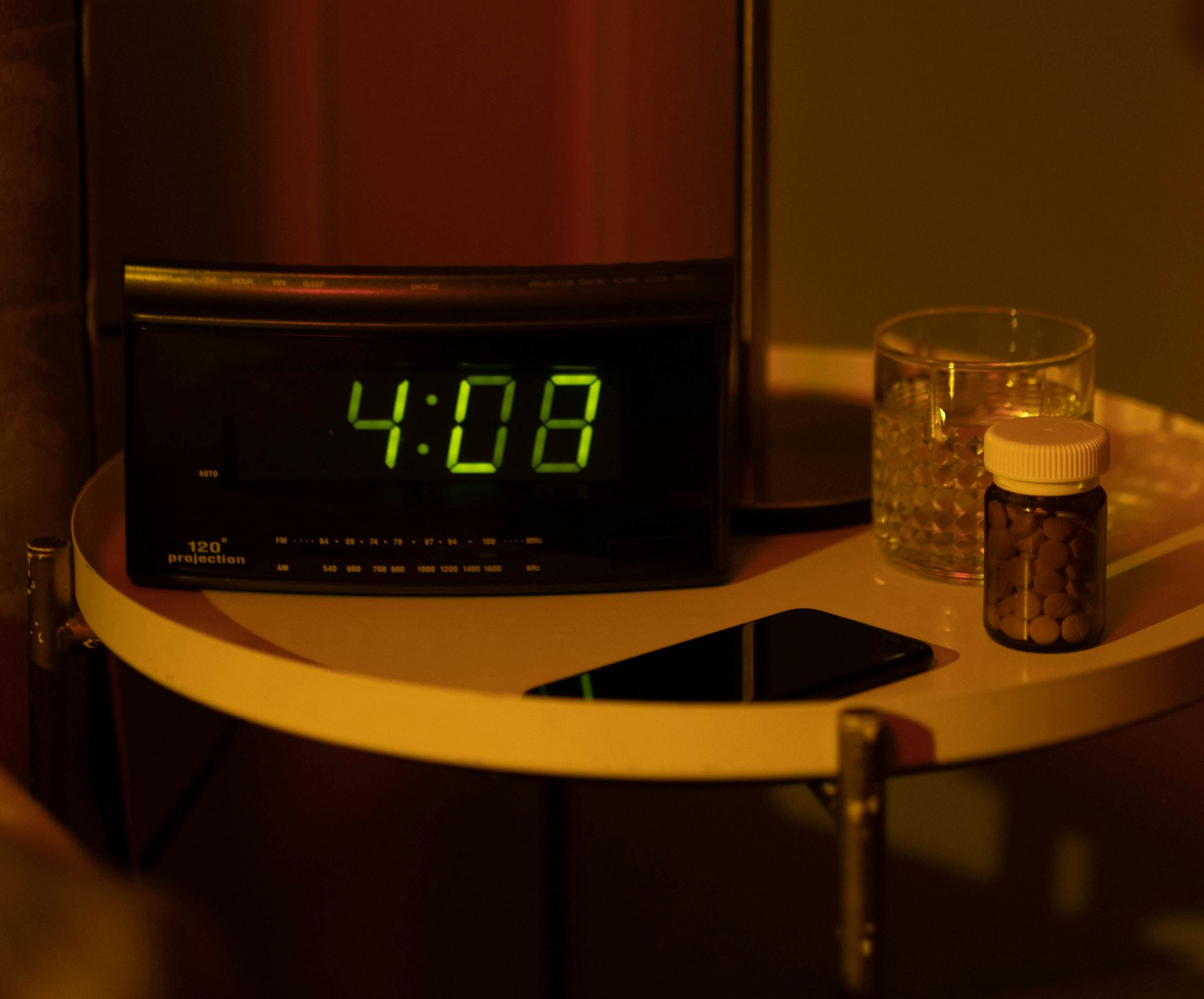 Digital clock showing 4:08 on a nightstand with a glass of water, medicine bottle, and a cell phone.