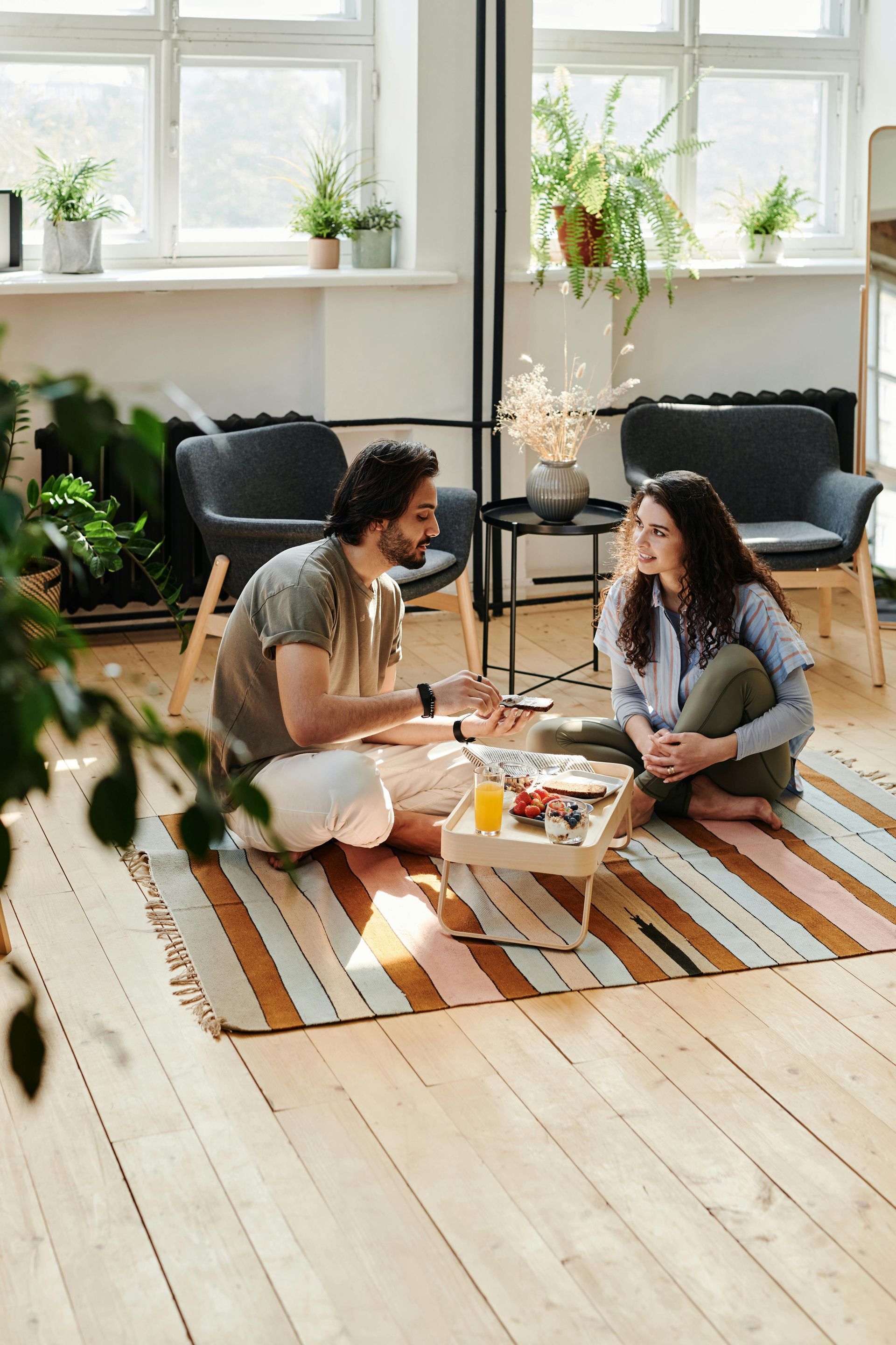 Couple sitting on rug in sunny room, eating and talking.
