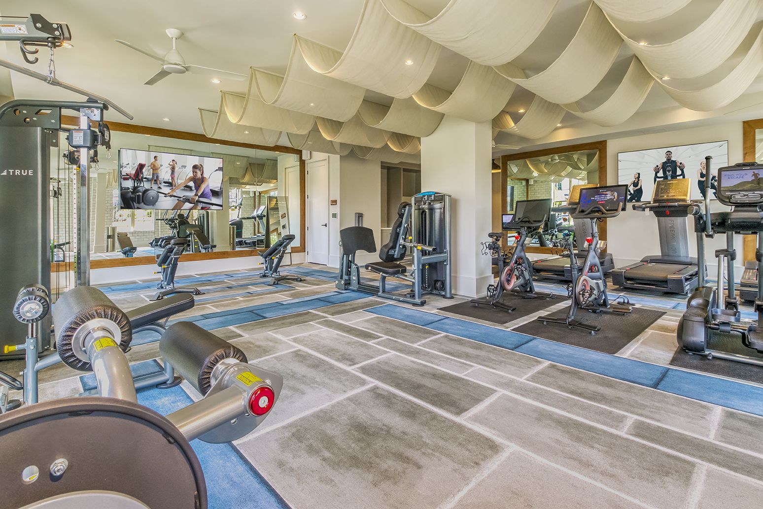 Gym with weight machines, dumbbells, and exercise balls. Large mirror reflects the interior.