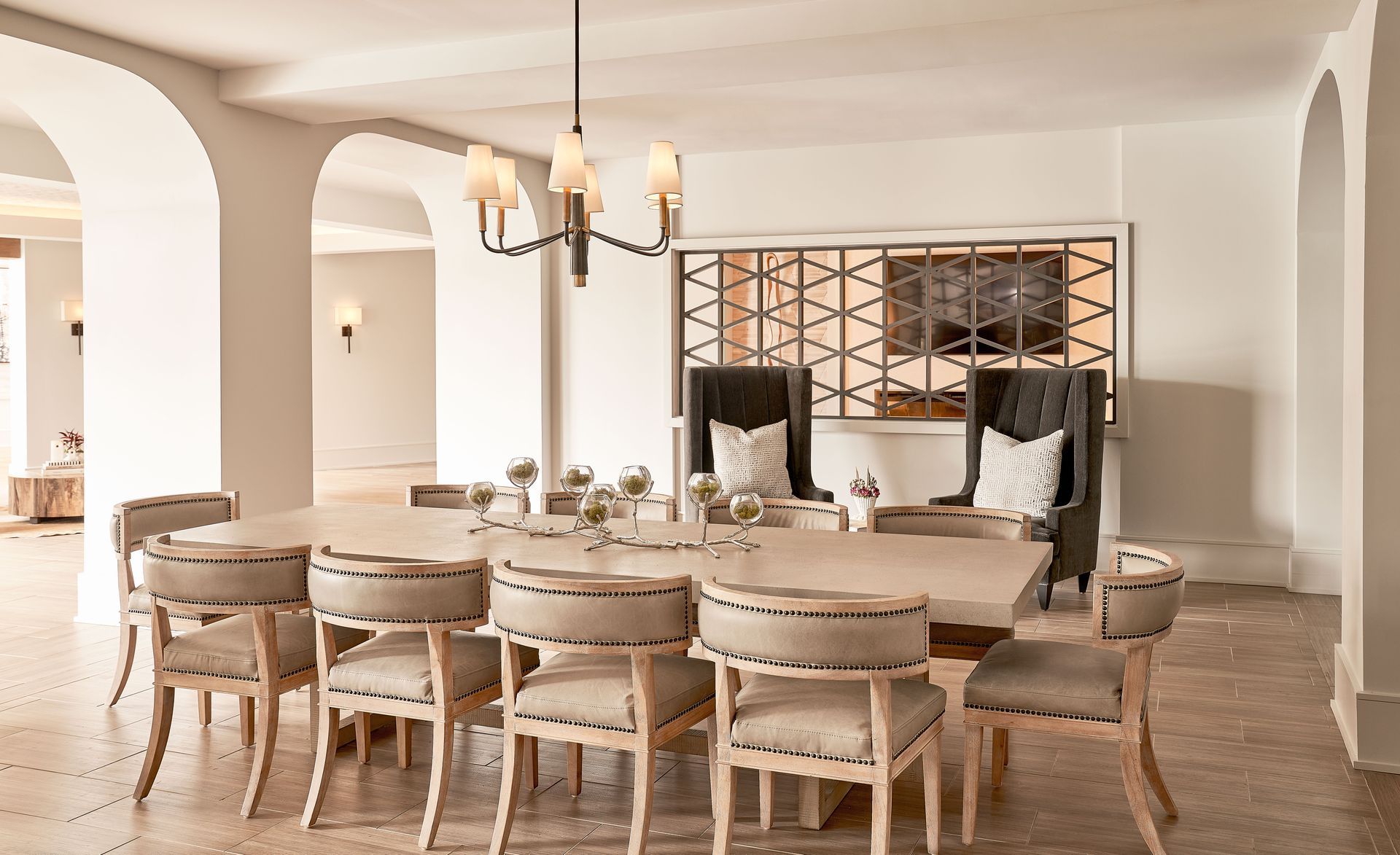 Elegant lounge with large table, chairs, chandelier, and decorative wall panel. Pale colors, archways.
