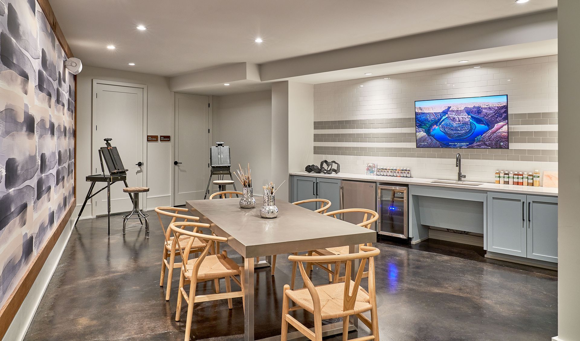 A modern room with a long table, chairs, art supplies, a TV, and a wet bar.