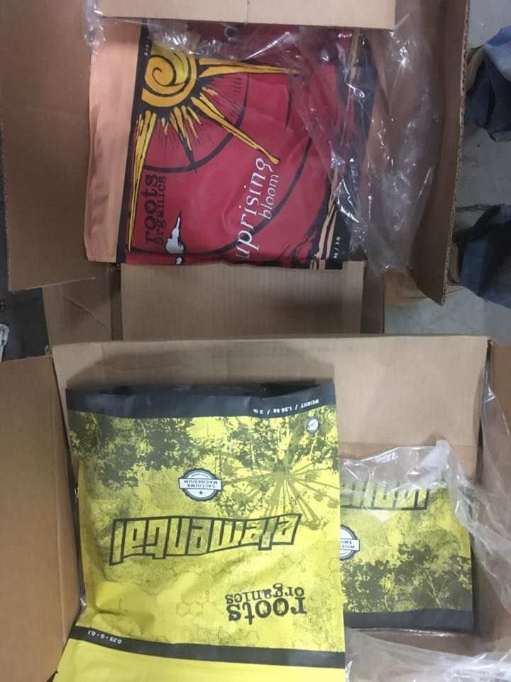 Boxes containing gardening supplies: soil bags (yellow) and a red Roots Organics bag.