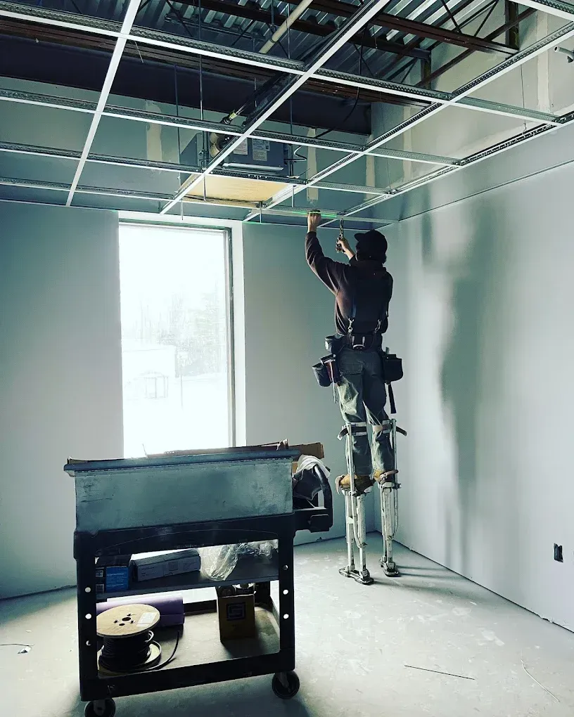 Construction worker on stilts installing a ceiling grid in a room with a window.