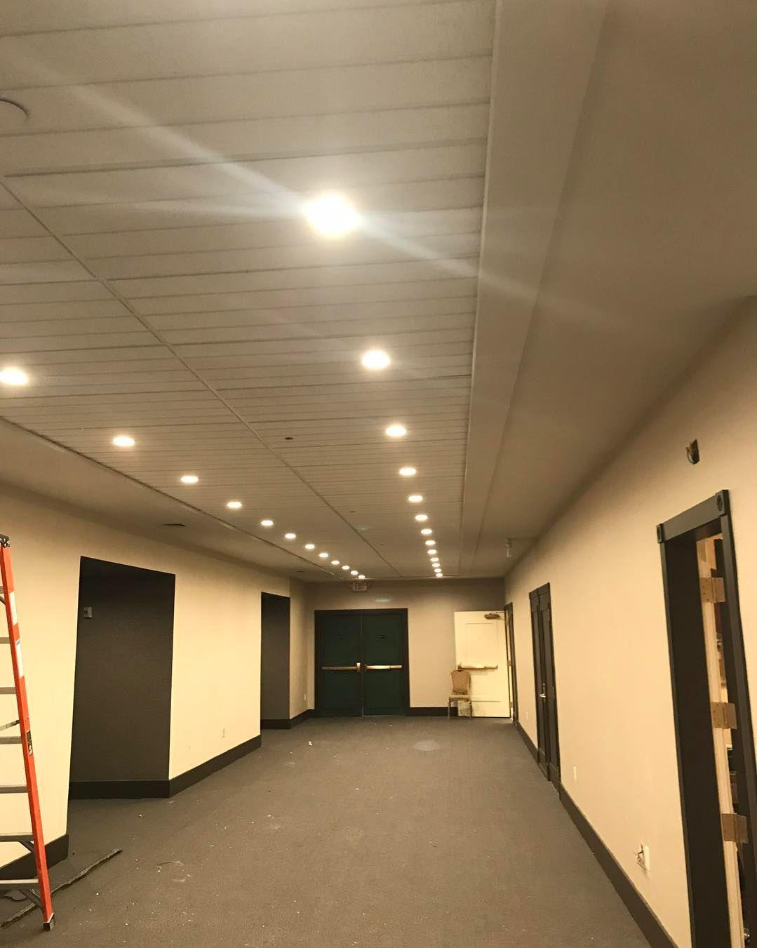 Hallway with recessed ceiling lights, tan walls, dark carpet, and black-framed doors.