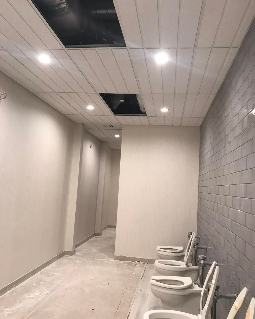 Public restroom with toilets, gray tile wall, and a drop ceiling with missing panels.