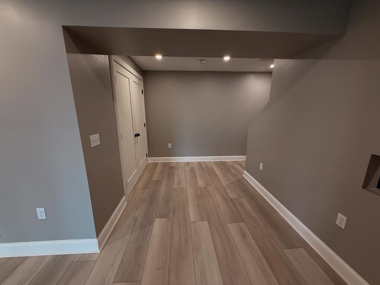 Gray-painted room with light wood-look flooring, white trim. Doorway leads to another room.