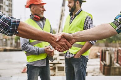 Two construction workers in high-visibility vests shake hands at a job site with unfinished buildings in the background.