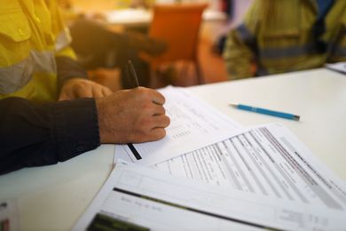A person in high-visibility work gear writes on a form at a desk with another person and a pen nearby.