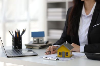 A professional sits at a desk with a laptop, stationery, and two house models, one yellow and one blue.