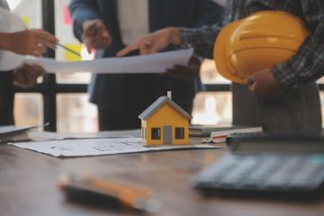 A miniature yellow house model on a table with a calculator, blueprints, and professional hands pointing at plans.