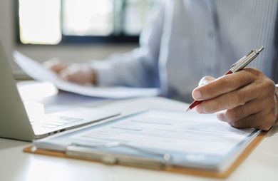 A person in a light shirt holding a red pen, filling out a document on a clipboard next to a laptop.