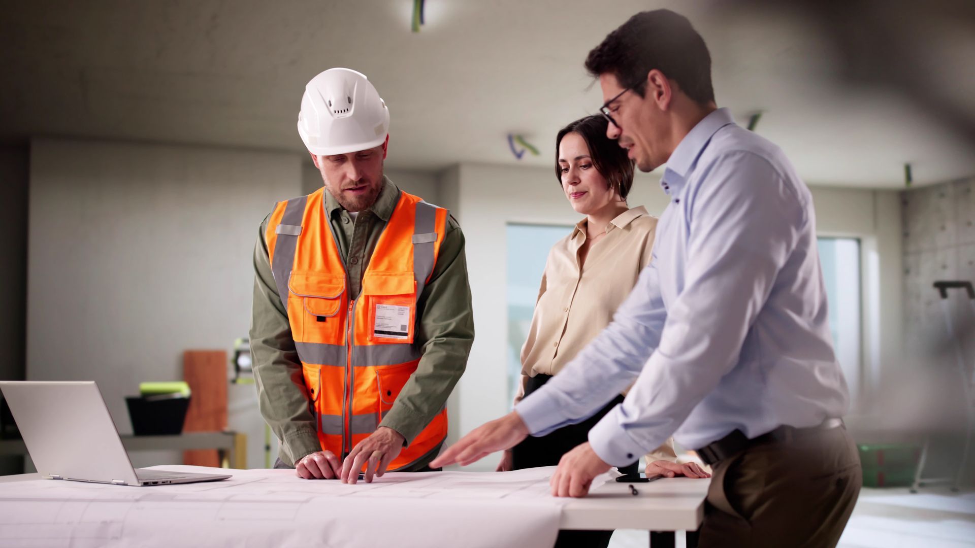 A construction supervisor and two colleagues in an indoor site office discuss blueprints on a table with a laptop.