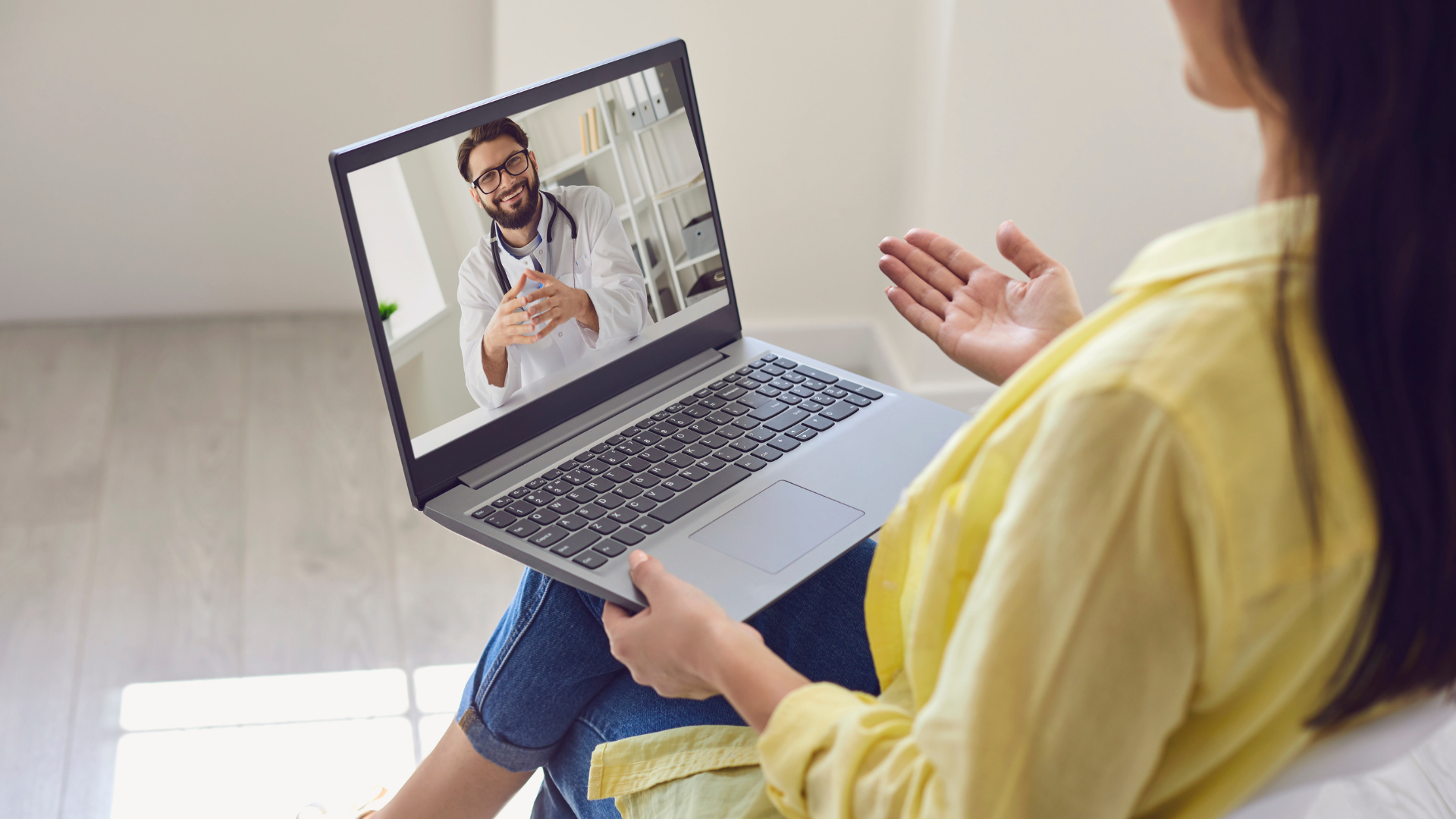 Woman in yellow shirt having a telehealth appointment with a smiling doctor on a laptop.