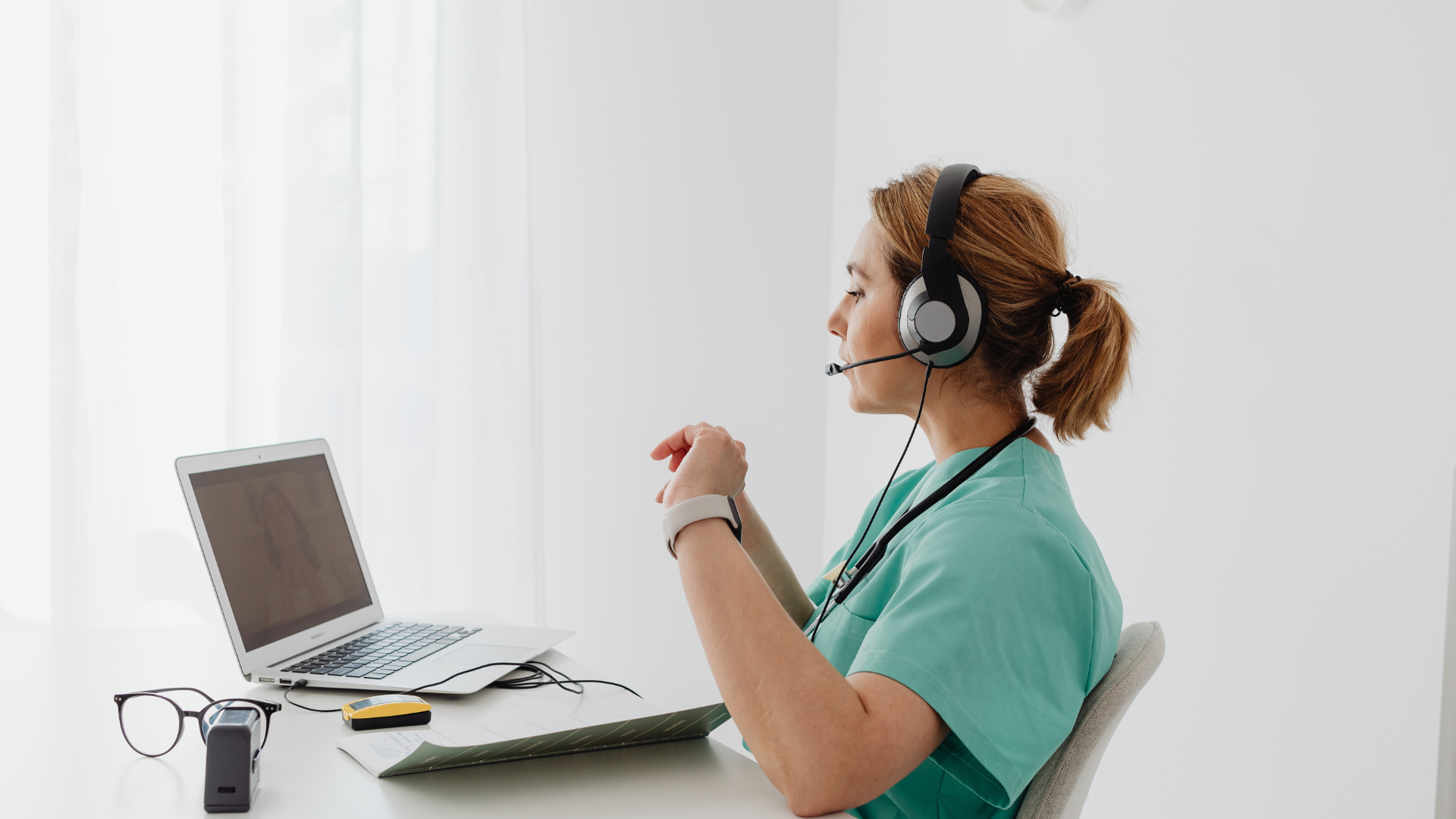 Medical professional wearing a headset, working on a laptop, in a bright room.