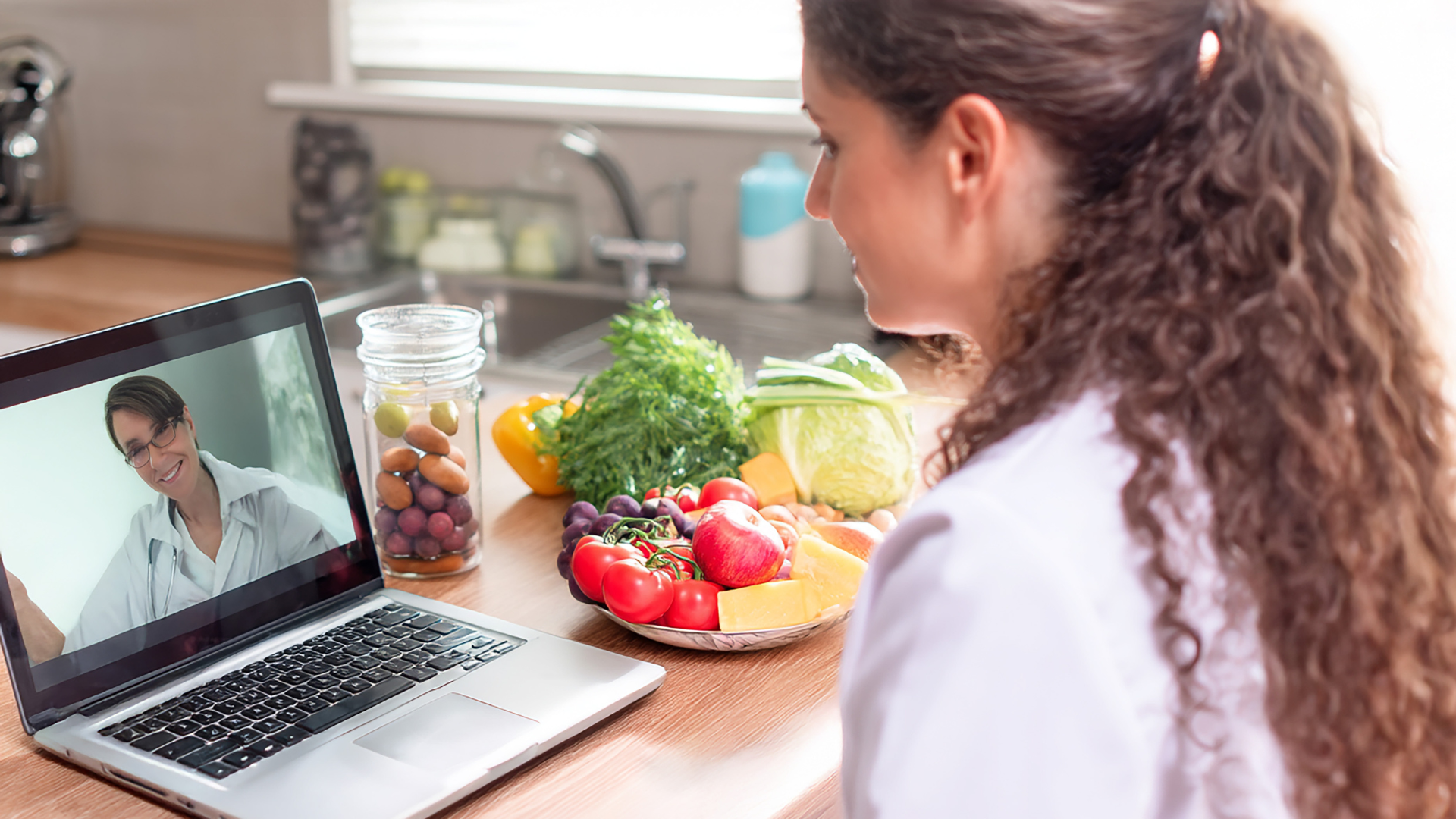 Woman in kitchen video chatting with doctor about fresh produce on counter.