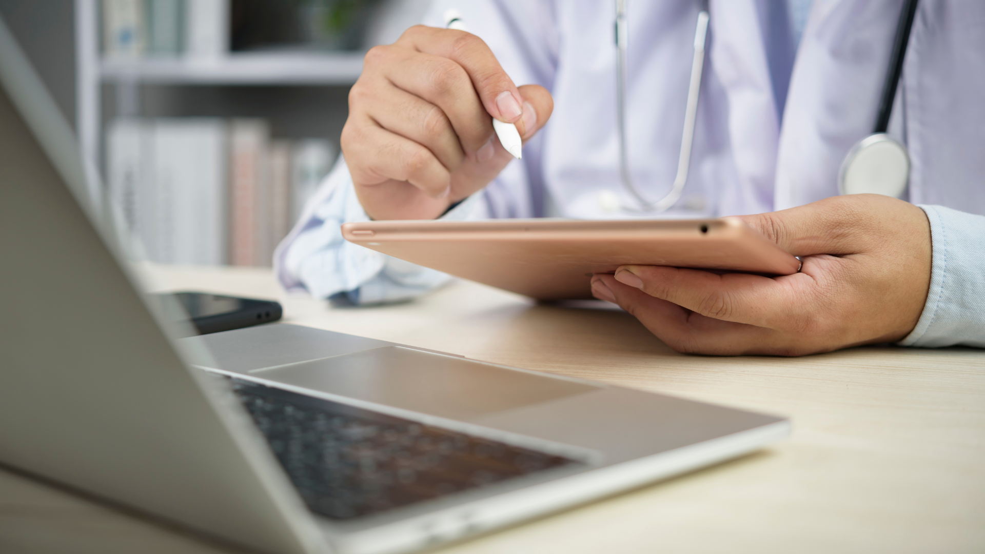 Doctor using a tablet and stylus, laptop in foreground.