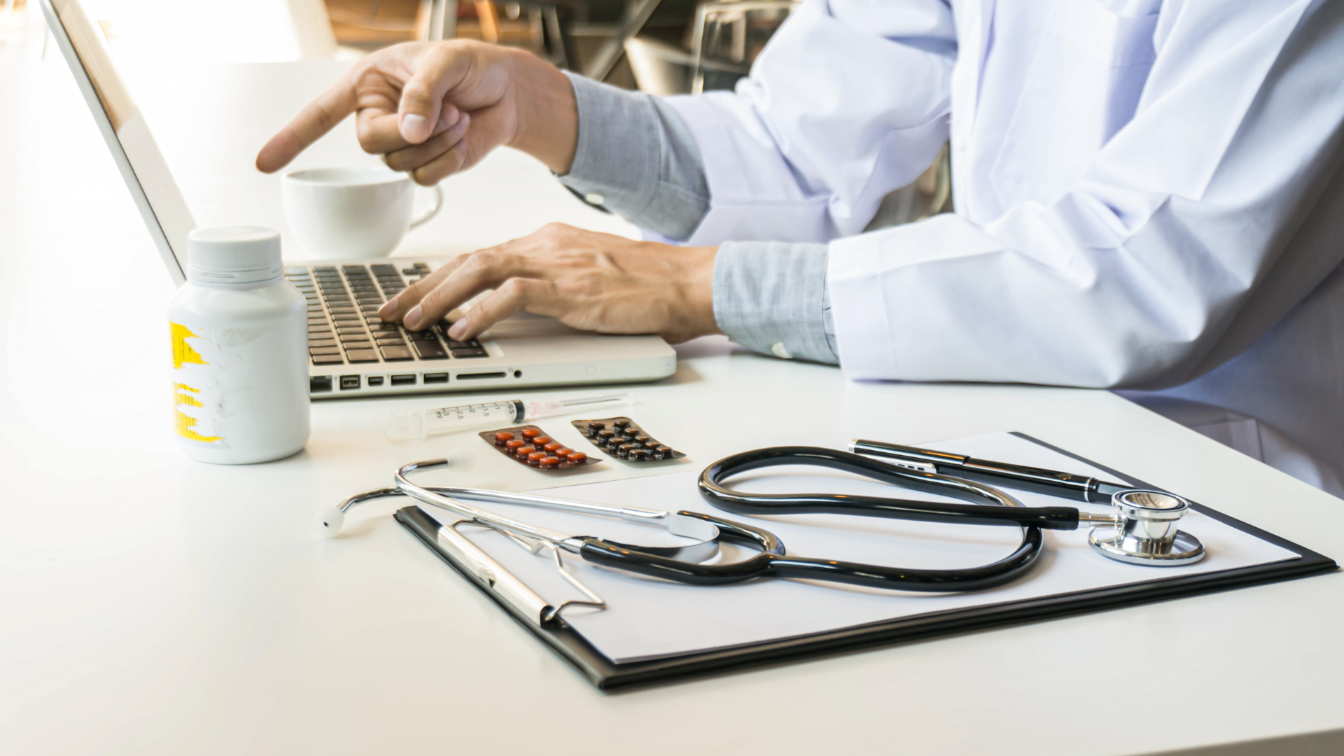 Doctor in white coat pointing at a laptop, stethoscope, and pills on desk.