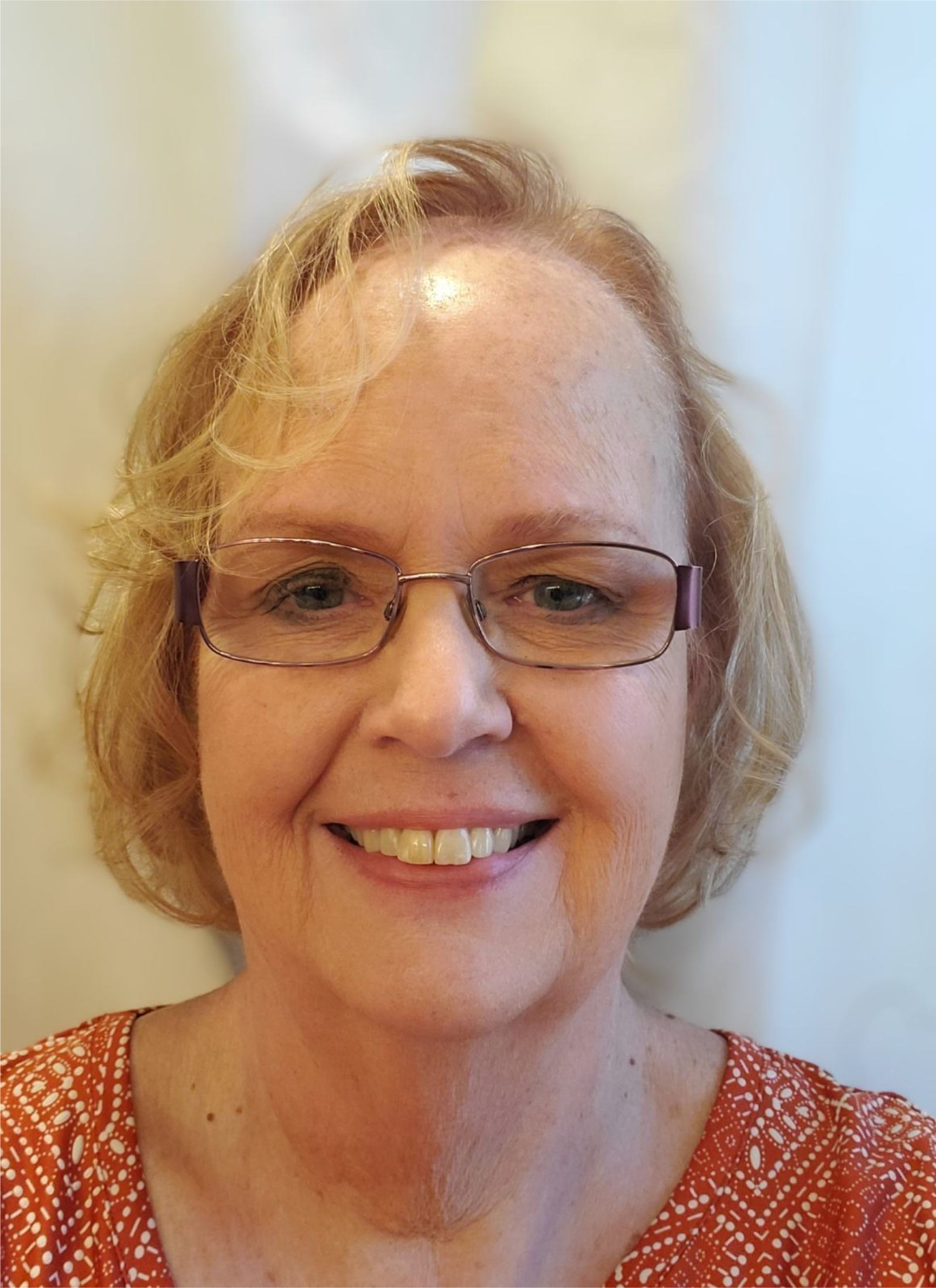 Woman with glasses smiles; blonde hair, orange patterned top.