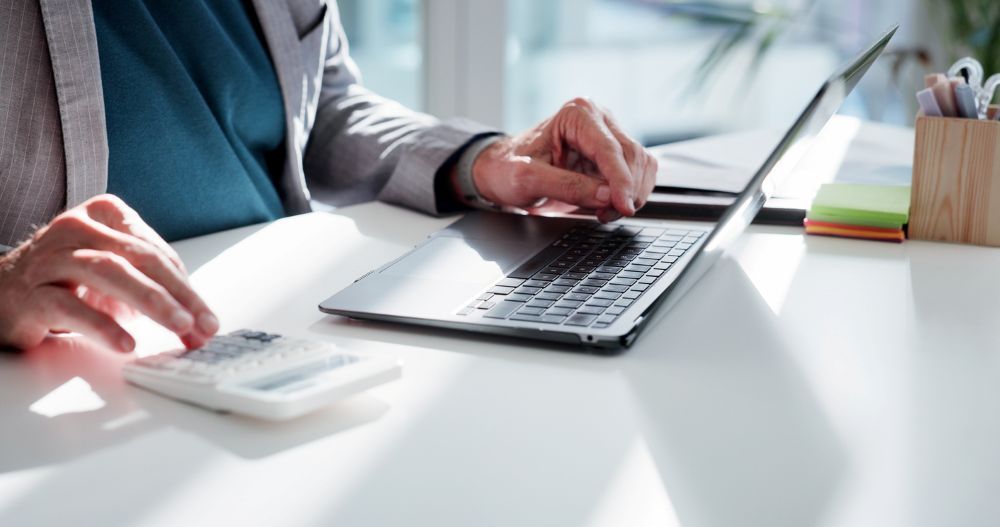 Man typing on a computer while using calculator representing payroll processing, financial analysis