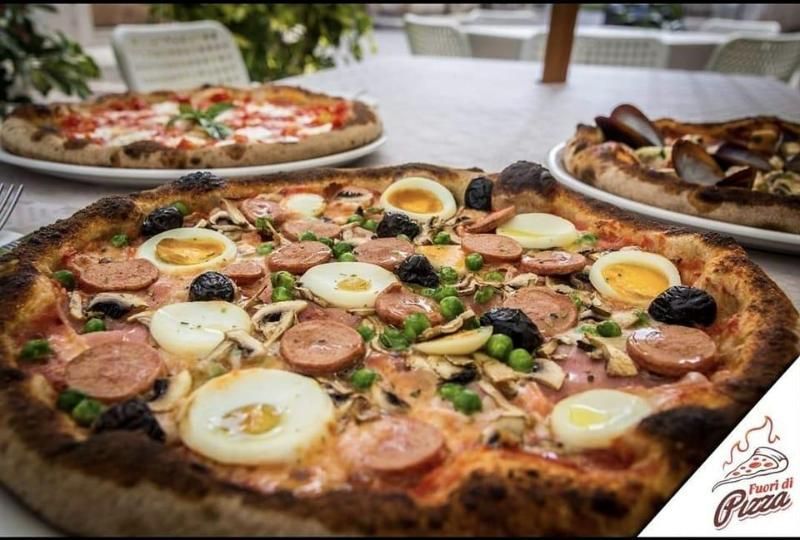 Su un tavolo ci sono tre pizze, di cui una in primo piano condita con salsiccia a fette, uova sode e piselli.