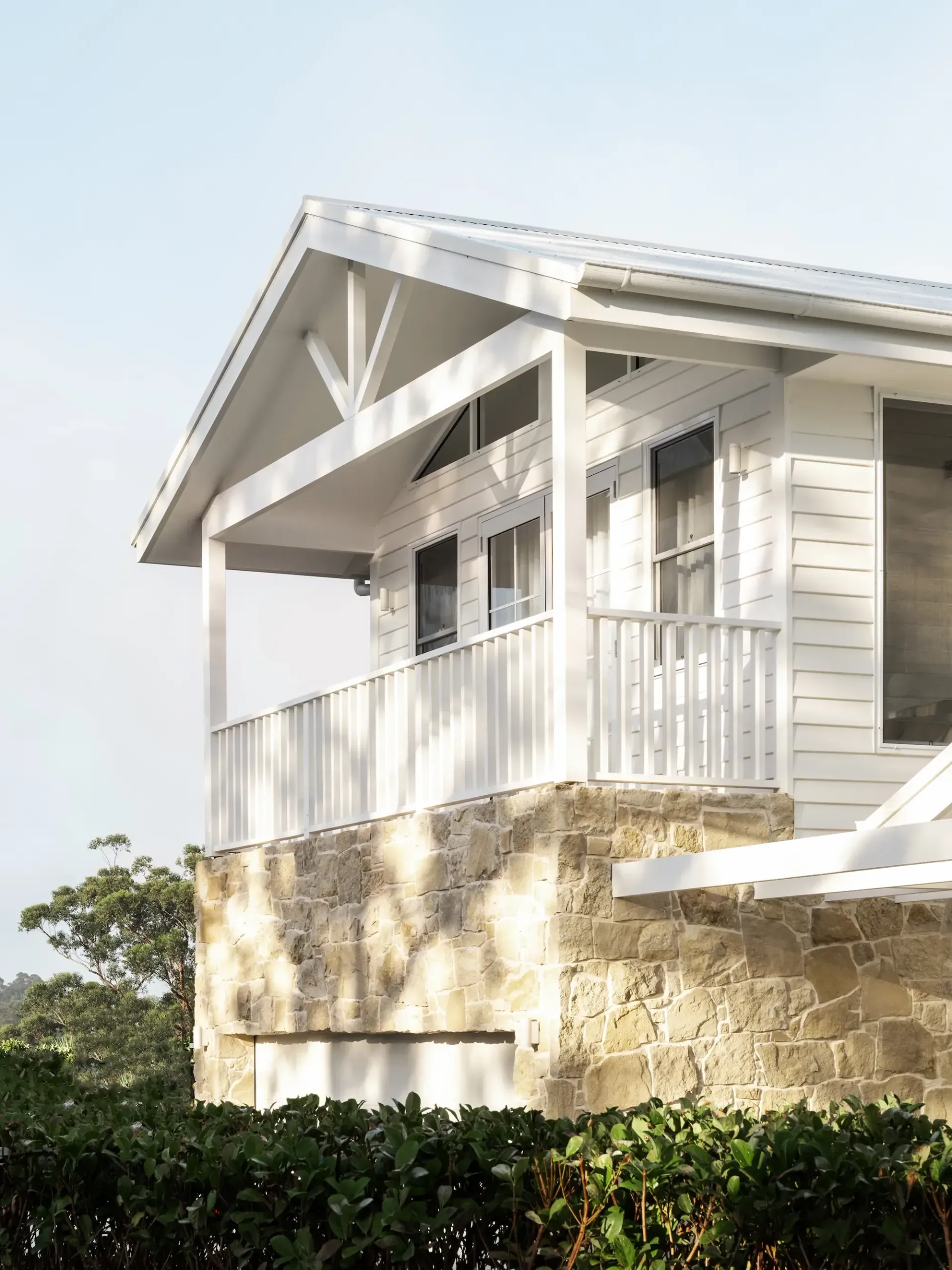 A white house with a gabled porch and balcony, set on a natural stone base with lush green hedges in the foreground.