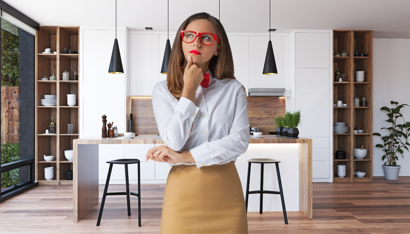 A woman wearing glasses is standing in a kitchen with her hand on her chin.
