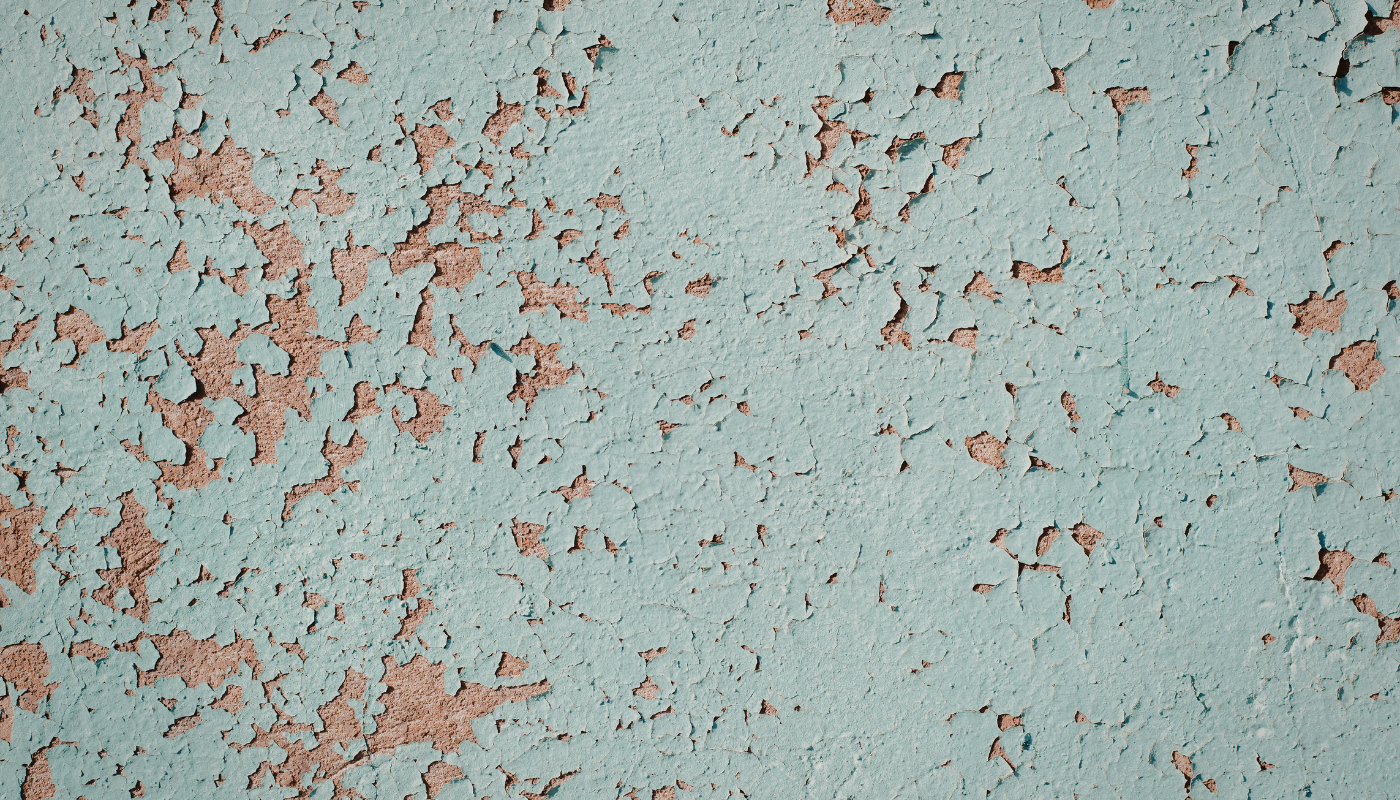 Close-up of a wall with peeling, faded blue paint revealing a tan-colored underlayer.