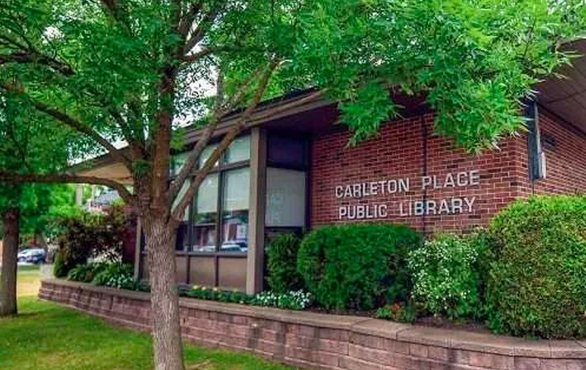Carleton Place Public Library building, brown brick facade, green bushes, and a tree on a sunny day.