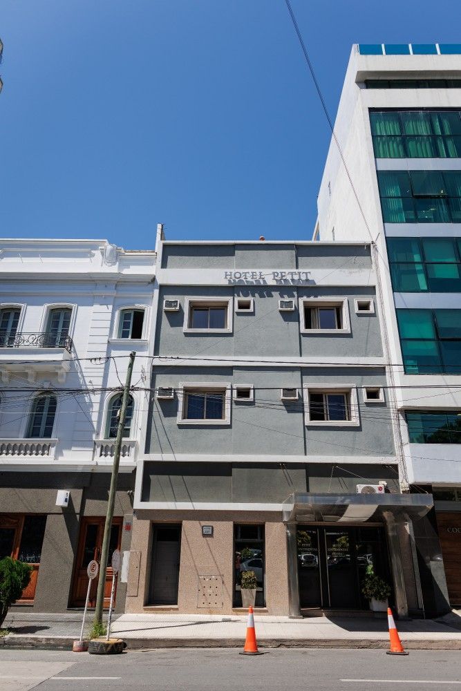 Edificio gris con pequeñas ventanas y un cartel, flanqueado por edificios blancos y modernos, bajo un cielo despejado.