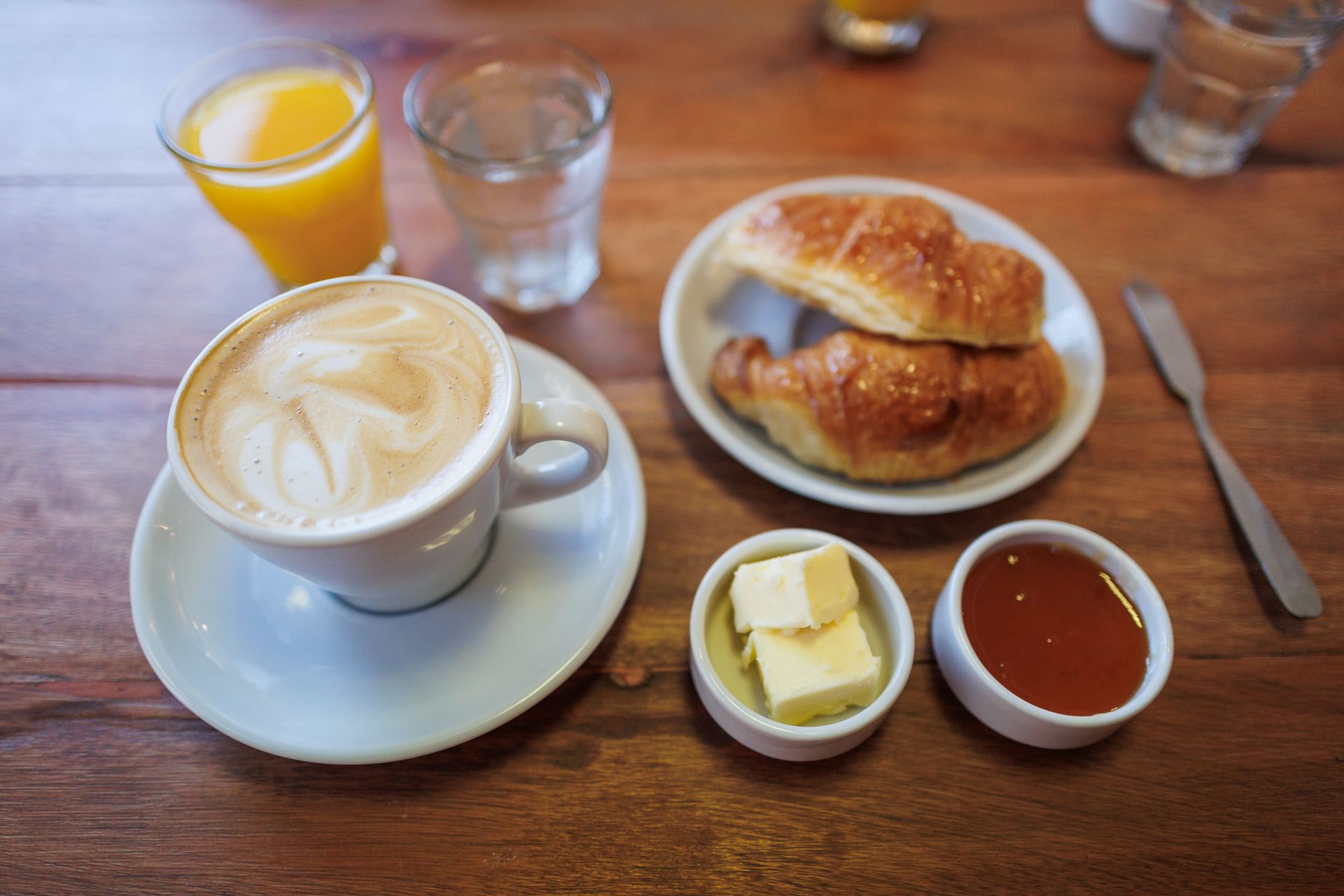 Desayuno: capuchino, croissants, zumo de naranja, mantequilla, mermelada y agua. Mesa de madera.