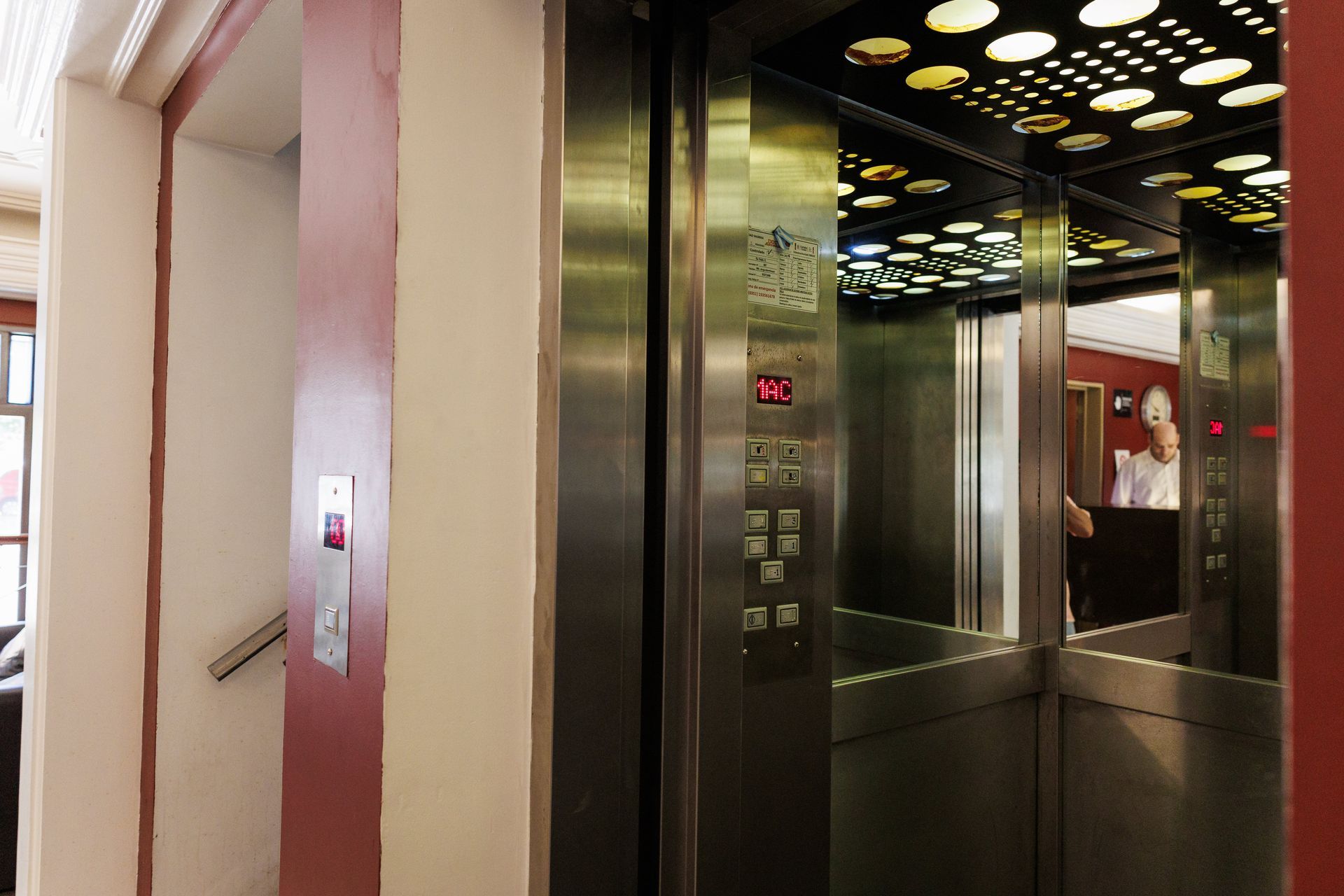 Ascensor con puertas abiertas, interior plateado. Botones e indicador de planta visibles. Entrada por pasillo.