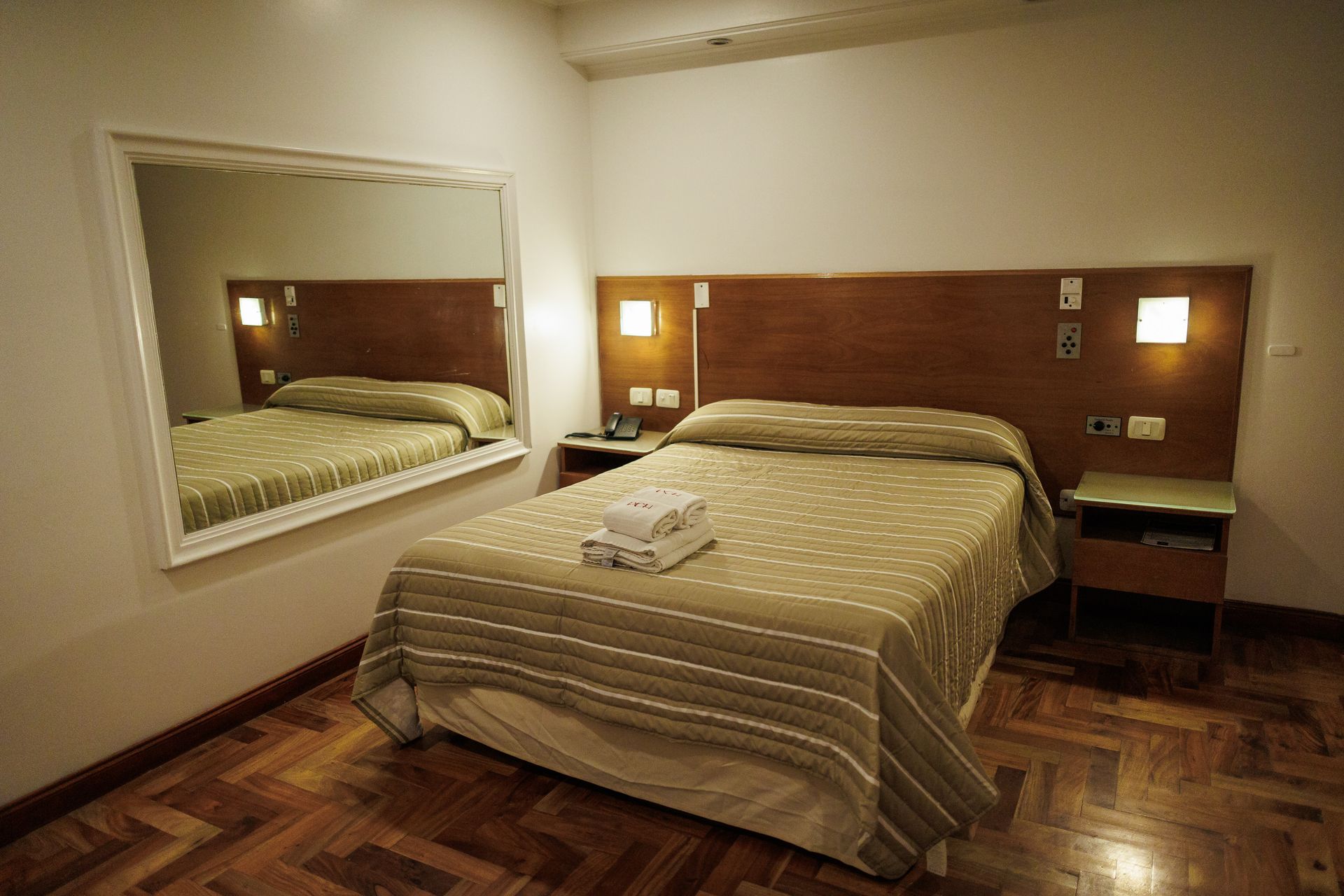 Habitación de hotel con cama, mesita de noche, espejo y suelo de madera.